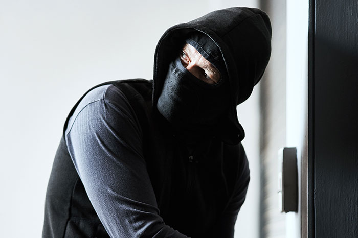 Masked man in a black hoodie cautiously peeking around a corner, representing wily criminals outsmarting police officers.
