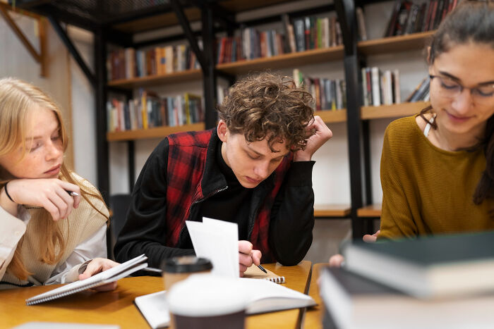 Three young people studying together in a library, focused on notes about most likely to succeed stories.