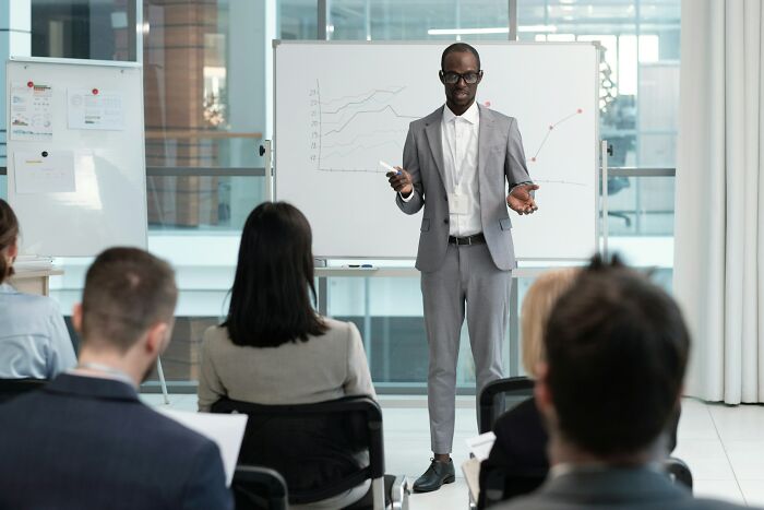 Man in a gray suit presenting to an audience, illustrating the story of the most likely to succeed with plot twists.