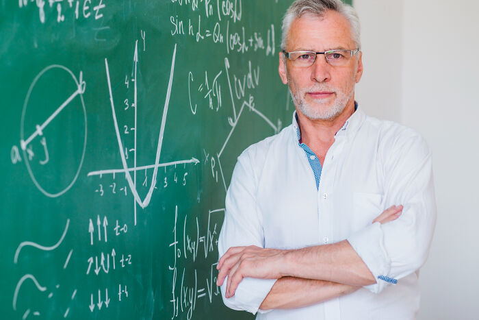 Mature male teacher with glasses standing confidently by a chalkboard filled with math formulas in a classroom.