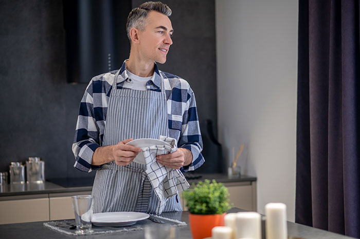 Man in a striped apron drying dishes at home, illustrating psychological cheat codes for social situations.