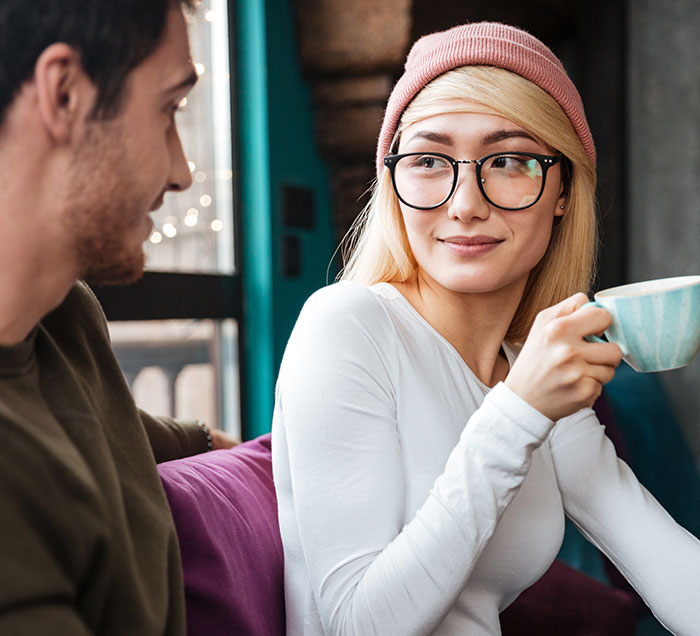 A young woman wearing glasses and a pink beanie holding a cup while talking to a man, illustrating psychological cheat codes.