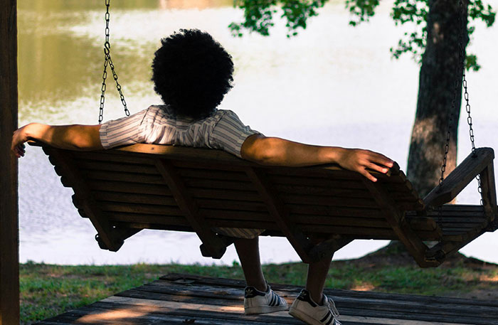 Person with curly hair relaxing on a wooden swing by the water, illustrating diabolical revenge stories concept.