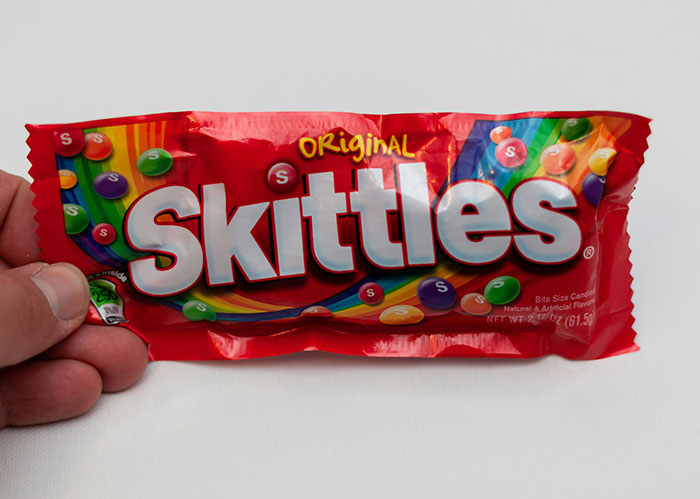 Hand holding a red Skittles candy pack with colorful sweets, illustrating diabolical revenge stories concept.