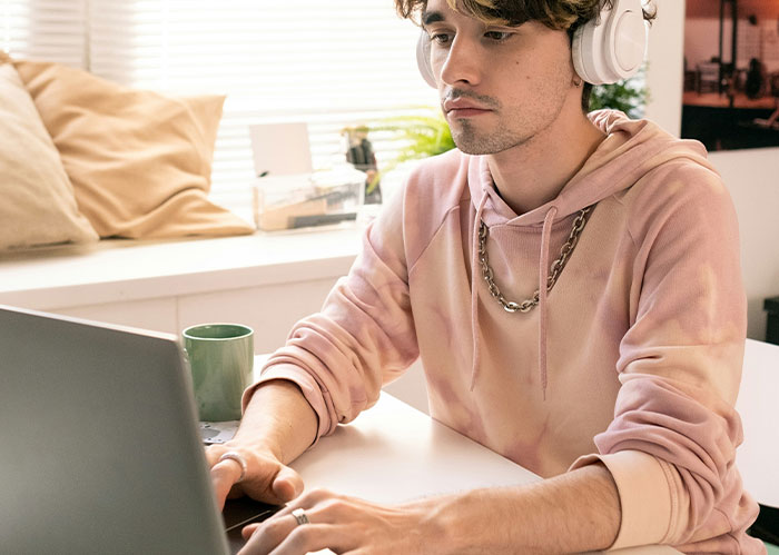 Young man wearing headphones, focused on laptop, creating or reading diabolical revenge stories at home desk.