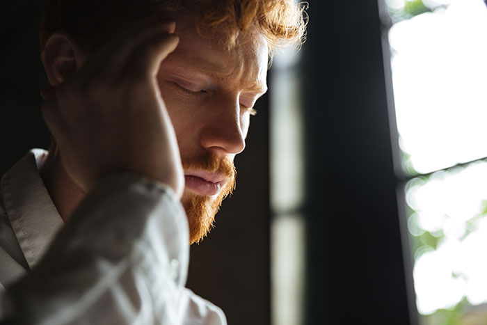 Red-haired man showing panic on his face, looking serious and deep in thought near a window.