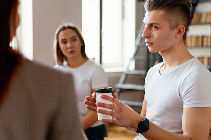 Young man with a panic expression holding a coffee cup, while others in the room scoff or look serious during discussion.