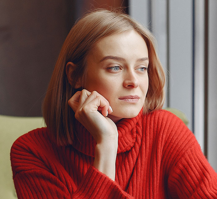Young woman in a red sweater looking thoughtfully out the window, reflecting on things people only believe once experienced