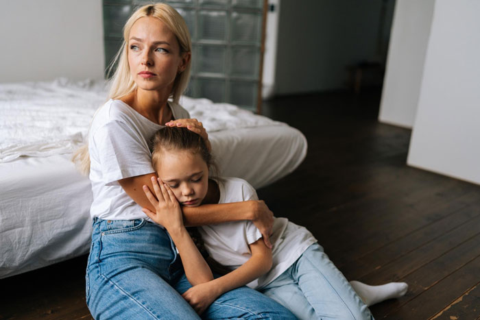Blonde woman comforting a sad child sitting on the floor, illustrating teen hostility towards stepmom in family setting.