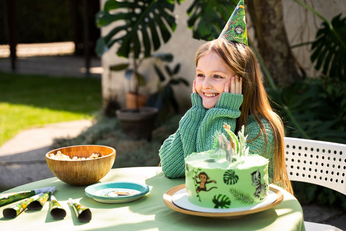 Teen at birthday party wearing a party hat, sitting next to a green jungle-themed cake and looking happy outdoors.