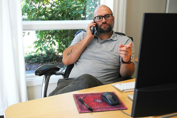 Man with glasses talking on phone and pointing, sitting in office chair by computer, workplace scandals concept.