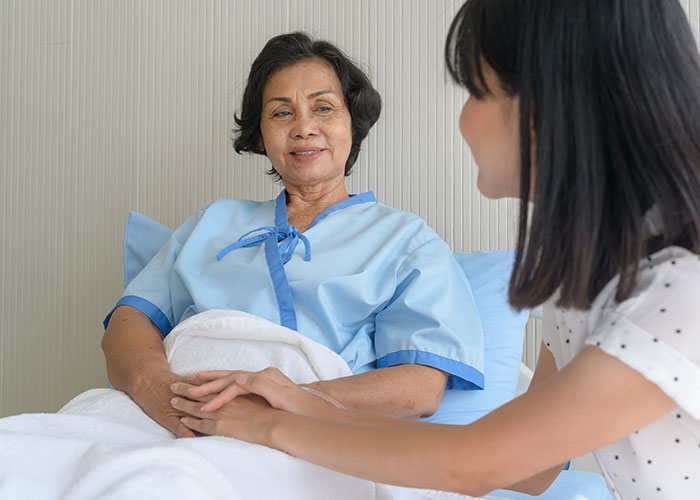 Patient in hospital gown resting in bed while a visitor holds her hands, highlighting post-anesthesia care moments.