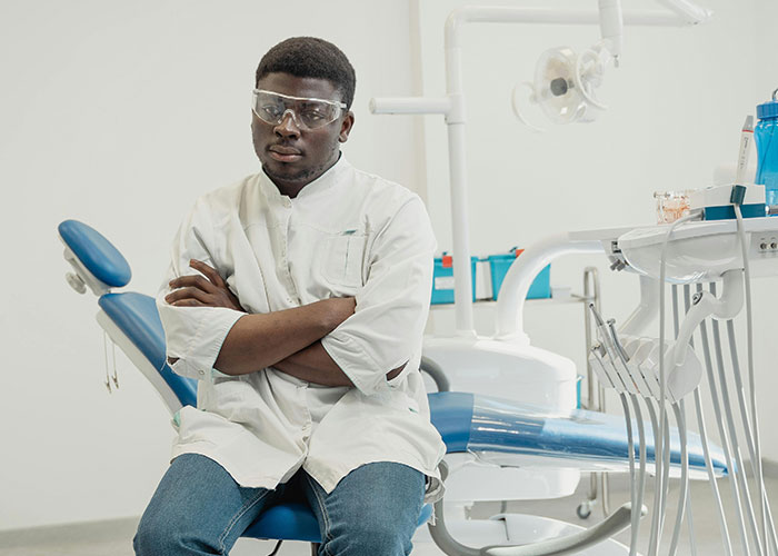 Young medical professional wearing protective eyewear sitting next to dental chair, highlighting post-anesthesia acts in healthcare.