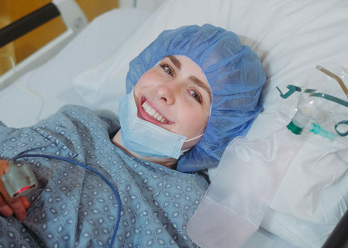 Patient smiling in hospital bed wearing surgical cap and mask, recovering with post-anesthesia care and monitoring equipment nearby