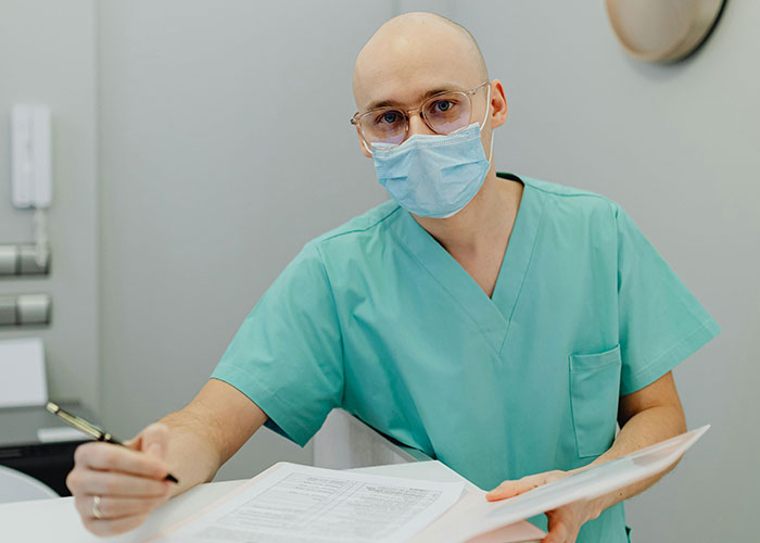 Doctor in green scrubs and face mask reviewing documents, related to wild and funny post-anesthesia acts in medical care.