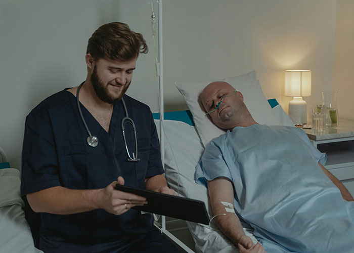 Doctor reviewing notes on tablet with patient recovering post-anesthesia in hospital bed, highlighting post-anesthesia acts.