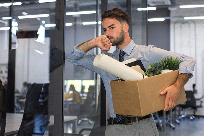 Young professional leaving office with box of belongings, showing subtle red flags indicating job insecurity.