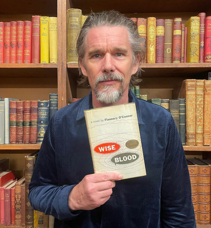 Ethan Hawke holding a book, pictured in front of a bookshelf, highlighting his role and online reaction pairing with Sydney Sweeney.