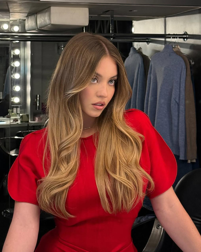 Sydney Sweeney showcasing natural beauty with styled hair and makeup in a dressing room setting with clothing in the background Sydney Sweeney showcasing natural beauty with styled hair and makeup in a dressing room setting with clothing in the background