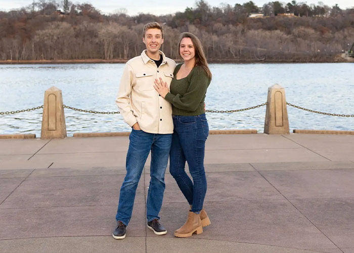 A smiling young couple standing by a river with fall trees in the background on a paved walkway.