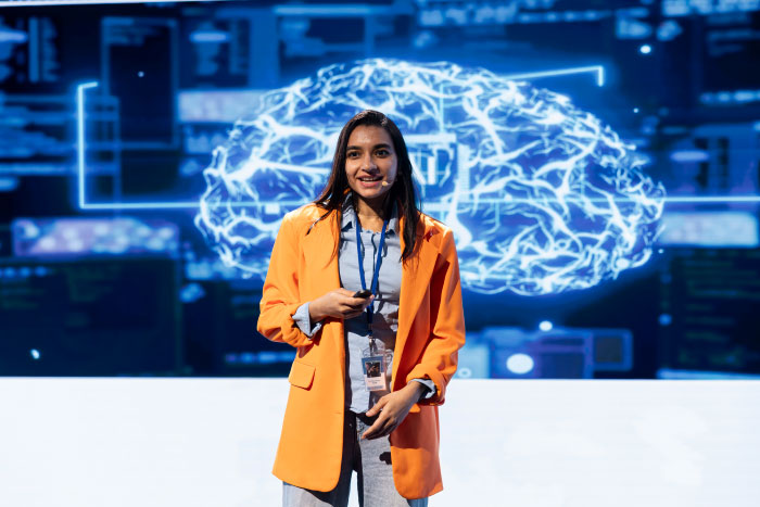 Woman in orange blazer presenting on stage with digital brain graphic in background, highlighting AI industry insights and secrets.