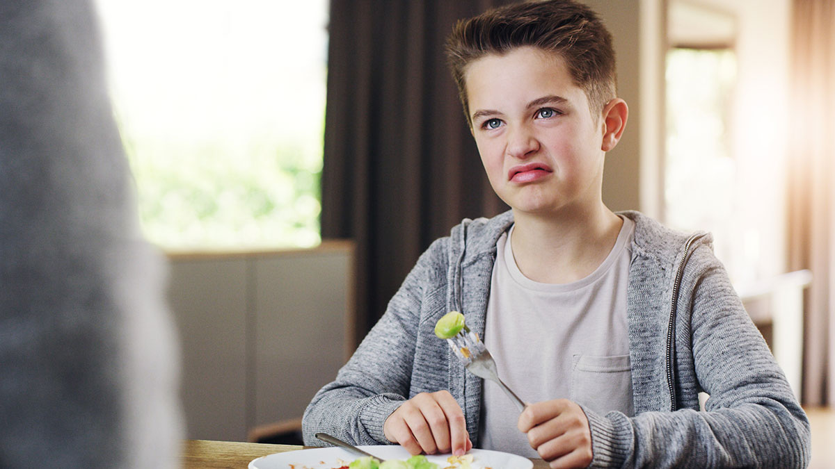 Teen making a disgusted face while holding a fork with onion during a picky meal shared with a cousin at home.
