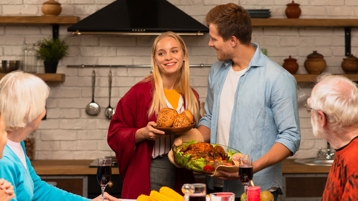 Young couple bringing Thanksgiving dinner to parents and friends at main table, son annoyed at overflow seating arrangement.