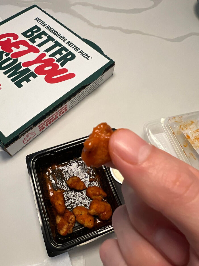 Close-up of a hand holding a small sauced chicken piece with a pizza box and container showing outrageous meals.