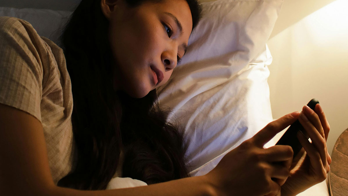 Young woman lying in bed using smartphone late at night, illustrating relatable habits many people are guilty of doing.