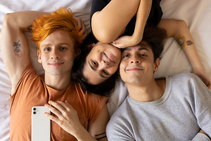 Three friends lying on a bed taking a selfie, representing must-try experiences that are overrated or overpriced.