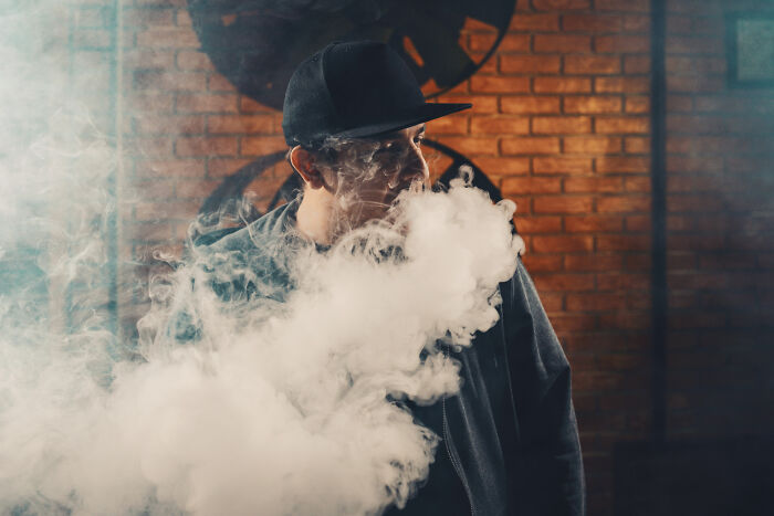 Young man wearing a black cap exhaling thick smoke in a dimly lit indoor space, illustrating overrated experiences.