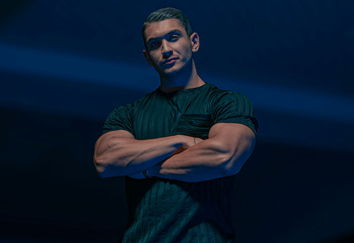 Muscular man with arms crossed, standing confidently in low light, representing female roasts hitting men hard.