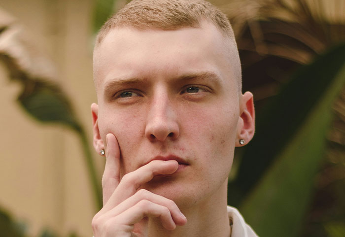 Young man with short hair and earrings, thoughtfully touching his face outdoors with blurred foliage background, female roasts.