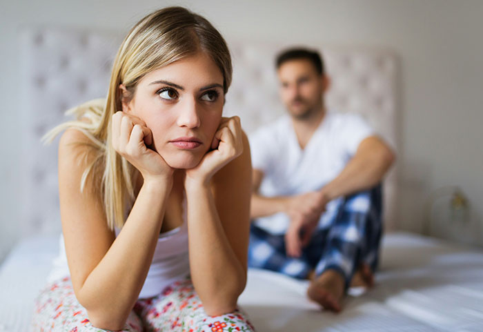 Young woman looking upset and thoughtful on bed while man sits in background, illustrating female roasts hitting men hard.