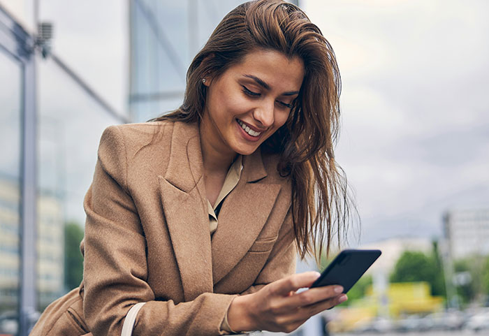 Young woman in a beige coat smiling while reading female roasts on her phone outdoors in an urban setting