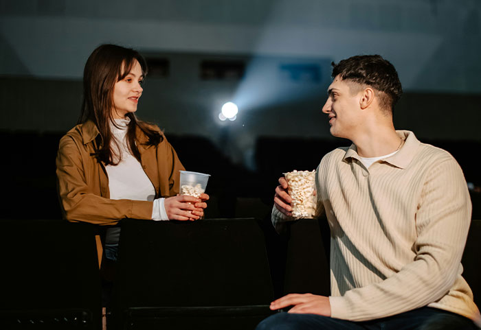 A woman and a man sitting in a theater holding popcorn, representing female roasts hitting men hard in conversation.
