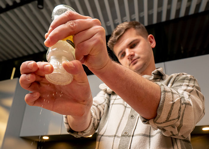 Young man salting a pot of chili in a kitchen, illustrating relationship conflict over food preparation and seasoning.