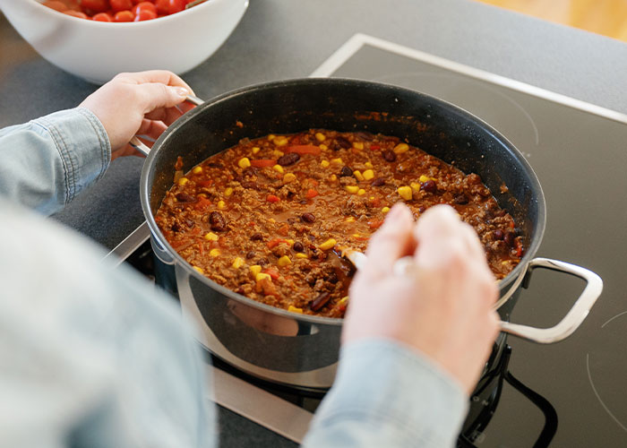 Person stirring a large pot of chili on stovetop with visible beans, corn, and ground meat ingredients. Person stirring a large pot of chili on stovetop with visible beans, corn, and ground meat ingredients.
