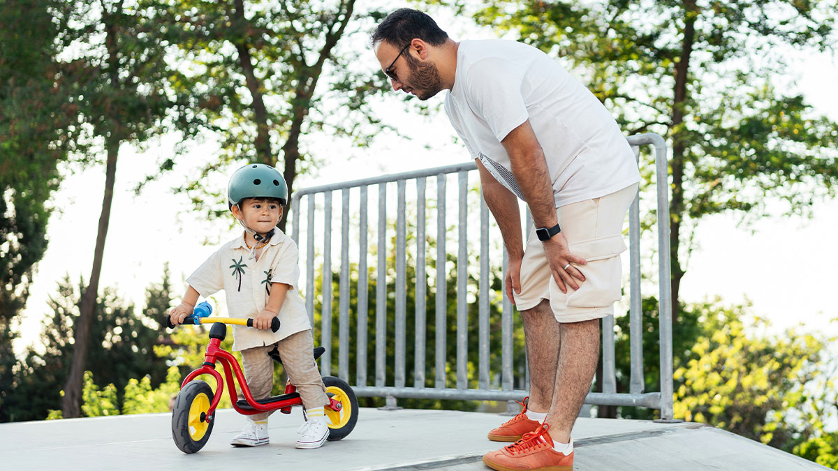 Father and son outdoors, young boy wearing helmet learning to ride bike, highlighting dad privileges in parenting moments.