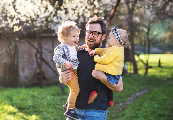 Father enjoying time outdoors holding two young children, highlighting dad privileges in parenting moments.