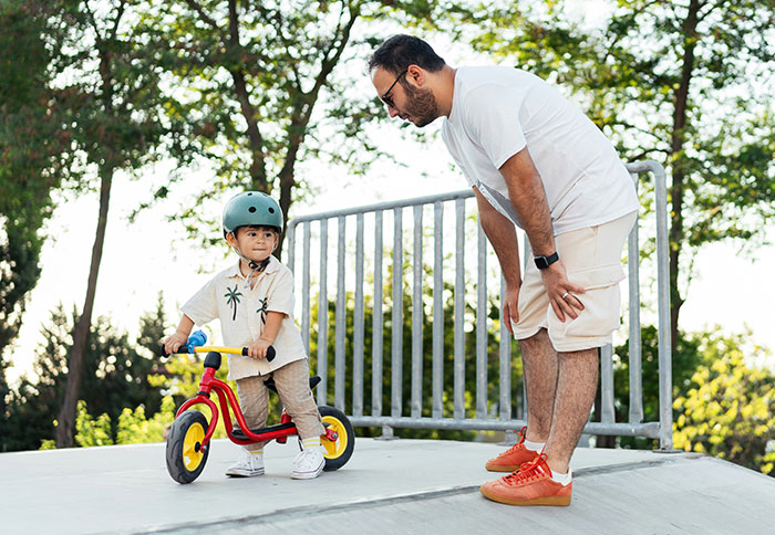 Man wearing glasses helping young child with helmet ride bike outdoors, illustrating dad privileges and parenting moments.