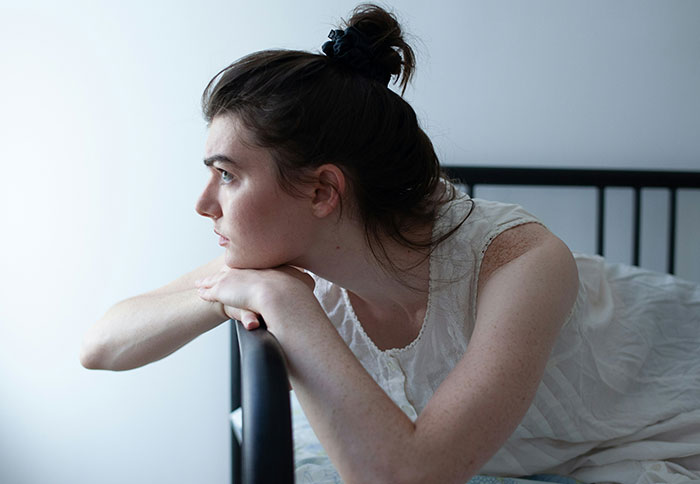 Woman in a white dress leaning on a bed frame, looking contemplative about dad privileges and parenting challenges.