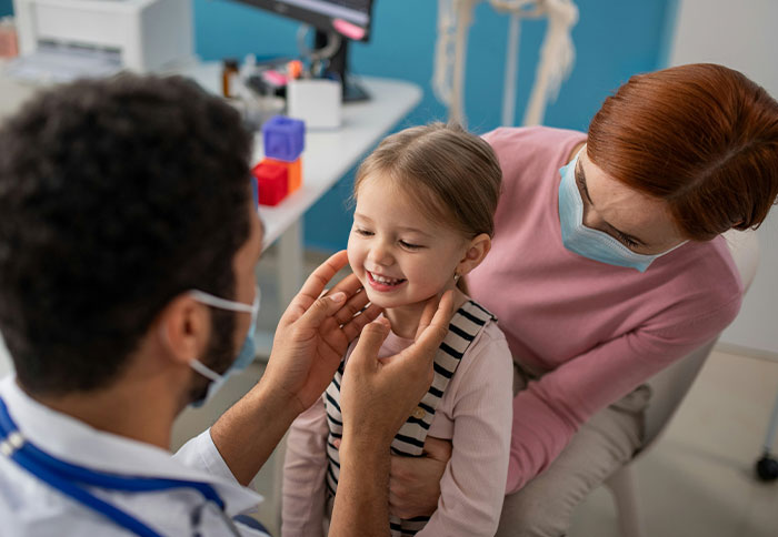 A doctor wearing a mask examines a smiling child held by a masked woman, highlighting dad privileges from moms' perspectives.