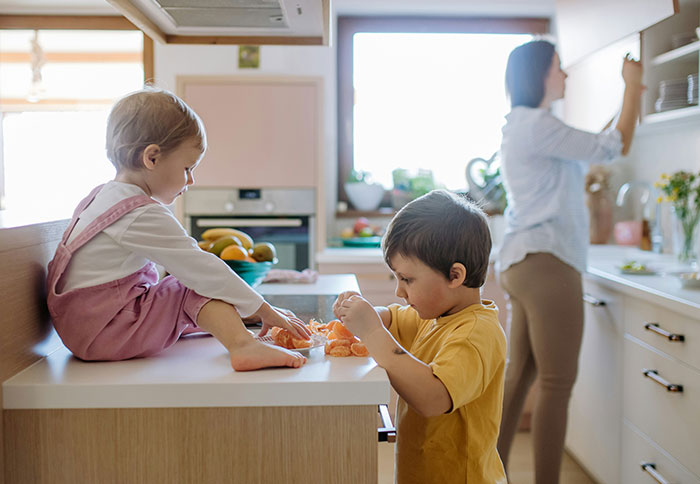 Two children peeling fruit in a kitchen while mom prepares food, illustrating mom and dad privileges in parenting dynamics.