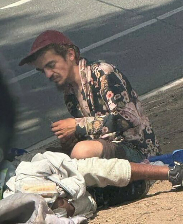 Man in floral jacket and cap sitting on street near struggling Nickelodeon star&rsquo;s motel stay scene outdoors.