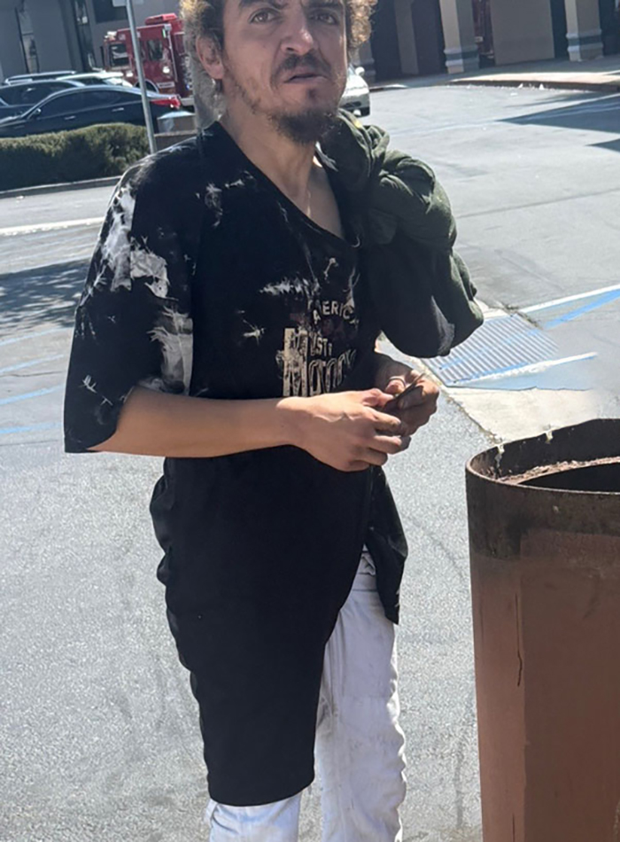 Nickelodeon star wearing a black tie-dye shirt and white pants, standing outdoors near a trash can in a parking lot.