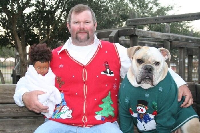 Man in a red Christmas sweater holding a doll next to a dog wearing a green holiday sweater in an awkward family Christmas photo.