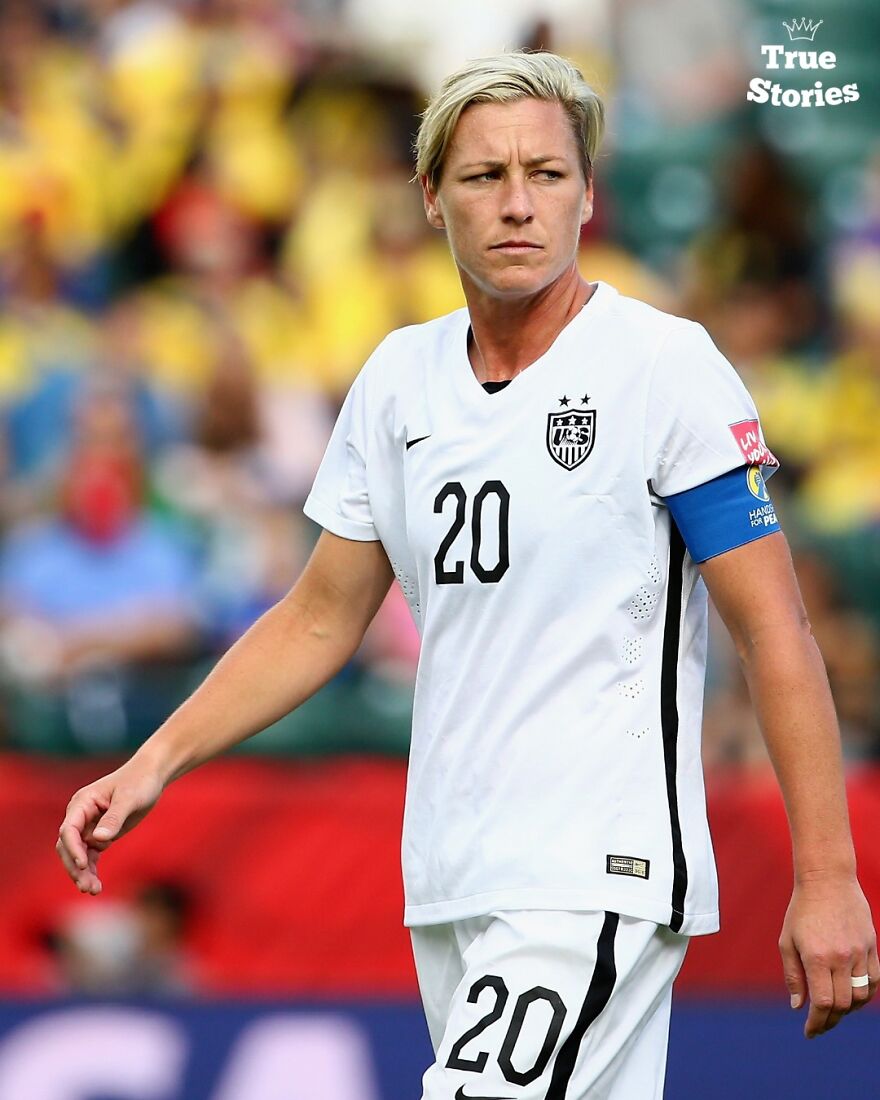 Female soccer player in white USA uniform with captain armband on field, representing important American personalities in sports.