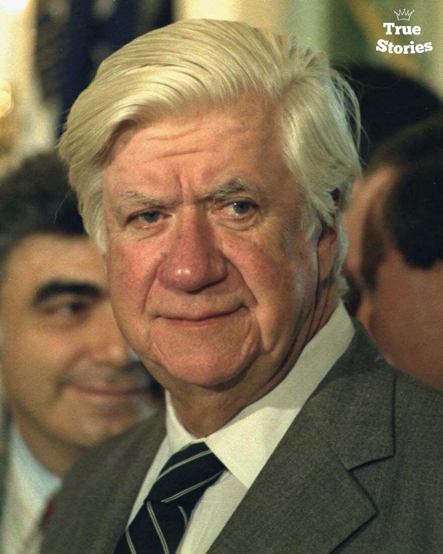 Elderly American man with white hair wearing a suit and tie among a group of important American personalities.