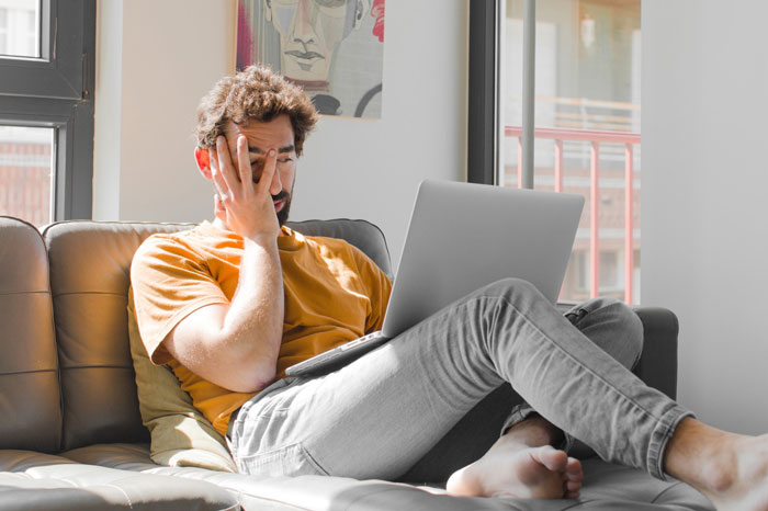 Unemployed husband sitting on couch with laptop, looking stressed about how to pay bills and manage finances.